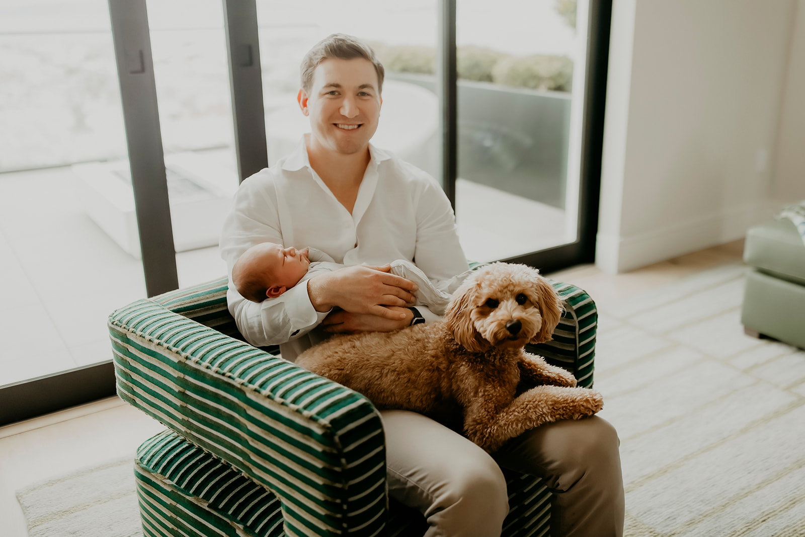 Shefali Parekh | Heartwarming In-Home Newborn Session: Maggie & Alex Black in Cow Hollow, San Francisco Man sitting on sofa holding his newborn with his dog